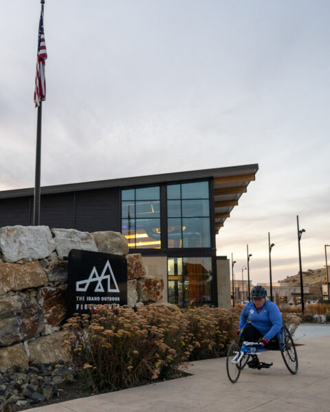 The image depicts a scene outside a modern building with large glass windows and a wooden overhang. The sky is overcast with soft, diffused lighting. In the foreground, a person wearing a blue jacket, helmet, and gloves is using a handcycle, moving along a paved path. To the left, an American flag is raised on a tall pole. Below it, a rock structure with a black sign displaying a logo is situated among dry, shrub-like plants. Sparse streetlights and barren hills can be seen in the distance.