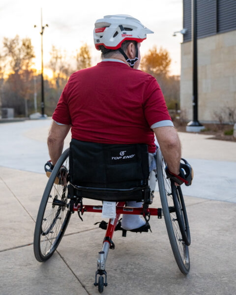 The image shows a person in a wheelchair viewed from behind on a paved outdoor path. The person is wearing a white helmet with grey and red accents and a red shirt with grey sleeves. They are using a sports wheelchair that has a black seat and a red frame. The path is smooth and leads out of view. The area is surrounded by trees with autumn foliage and lit by street lamps. On the right, there is a building with light-colored walls.