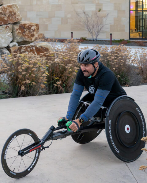 The image shows a person using a handcycle outdoors. They are wearing a black helmet, black gloves with green details, and a black and blue long-sleeve shirt. The handcycle has large, sleek wheels with the brand name "CORIMA" visible, and the frame is dark with the text "carbonbike" on it. The person appears focused and is pushing the pedals with both hands. In the background, there are large rocks, ornamental grasses, and a modern building with a beige stone facade. Some windows in the building are illuminated.