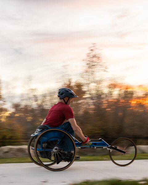 The image shows a person in a red shirt and blue helmet using a racing wheelchair on a smooth path. The scene captures a motion blur effect, suggesting speed and movement. The wheelchair is blue with two large rear wheels and a smaller front wheel. The background consists of a blurred view of trees and vegetation, illuminated by a soft, pastel-colored sky during sunset or sunrise, featuring hues of orange, pink, and light purple.