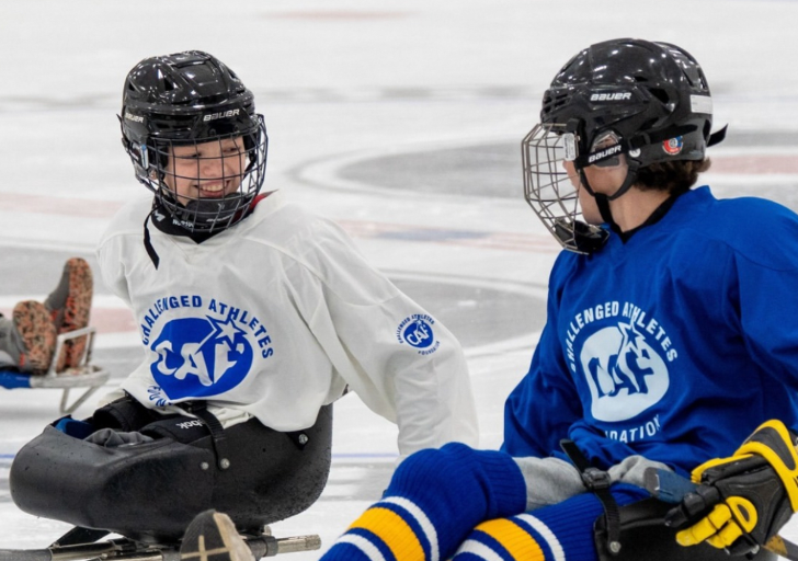 Two athletes with physical disabilities sit in sleds on the ice, smiling and talking while wearing CAF sled hockey jerseys during a clinic session.