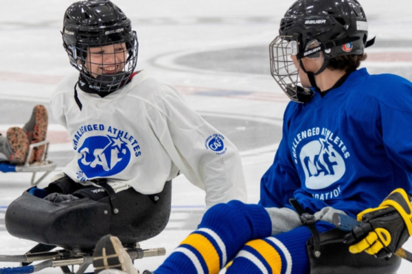Two athletes with physical disabilities sit in sleds on the ice, smiling and talking while wearing CAF sled hockey jerseys during a clinic session.