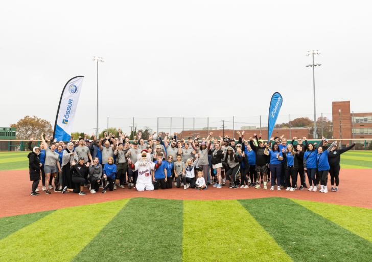 A large group of athletes with physical disabilities, coaches, volunteers, and partners stand together on a turf field, raising their arms and smiling during a CAF running and mobility clinic with Össur banners in the background.
