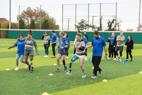 A group of athletes with physical disabilities walk together through a mobility drill on a turf field, supported by coaches and volunteers during a CAF running and mobility clinic.