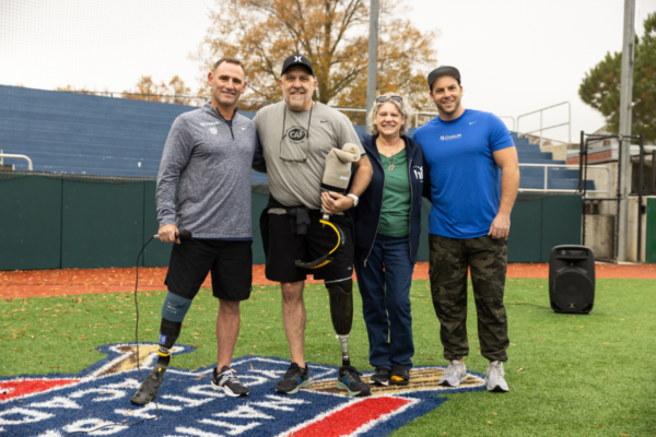 Four adult participants with lower-limb prostheses and clinic partners stand side by side on a turf field, smiling for a group photo at a CAF running and mobility clinic.