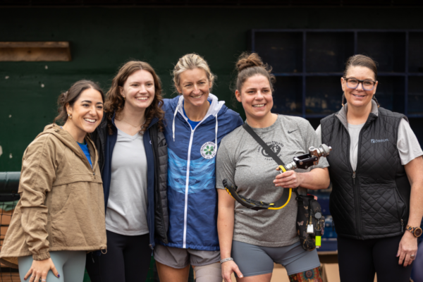 Five women stand shoulder to shoulder smiling at a CAF running and mobility clinic, including athletes with physical disabilities holding prosthetic components and wearing clinic apparel.