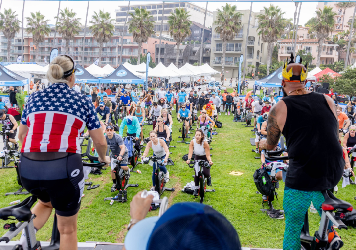A group of athletes and instructors ride stationary bikes on an outdoor stage during a Tour de Cove cycling class, wearing themed costumes while leading a large crowd at a beachfront event.