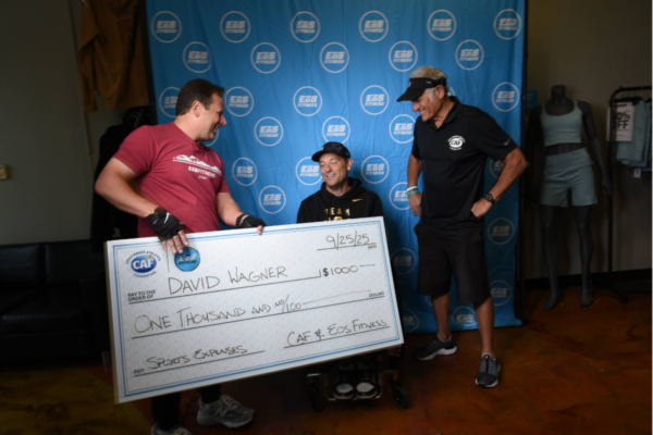 An athlete with a physical disability receives a CAF grant check inside an EōS Fitness location, standing alongside EōS staff and CAF leadership in front of a branded backdrop.