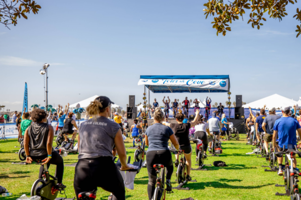Participants raise their arms while riding stationary bikes during an outdoor Tour de Cove cycling class, with instructors leading from the stage and spectators gathered nearby.