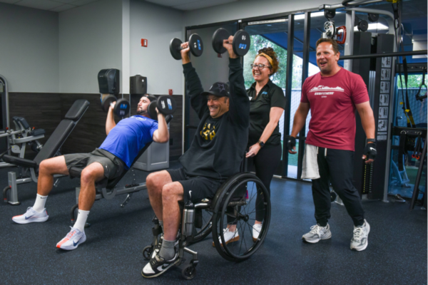 An athlete with a physical disability performs a seated dumbbell shoulder press in a wheelchair at an EōS Fitness gym, supported by trainers and staff during a strength training session.