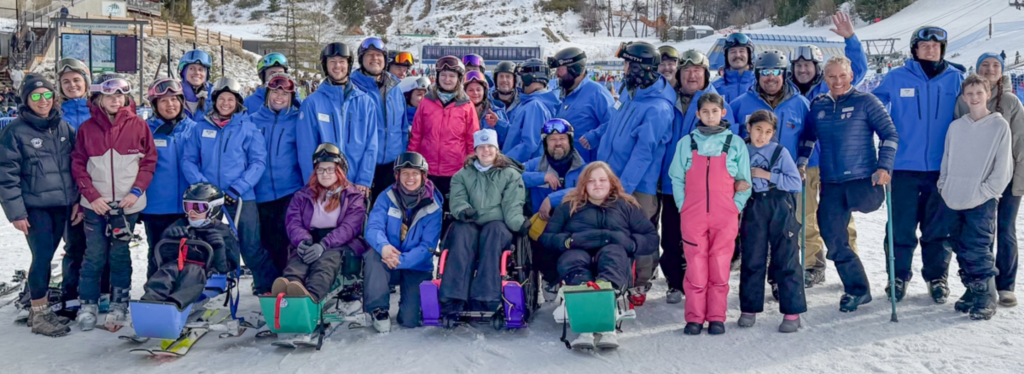 A group of people in ski gear, some using adaptive ski equipment, posing on a snowy slope with mountains in the background.