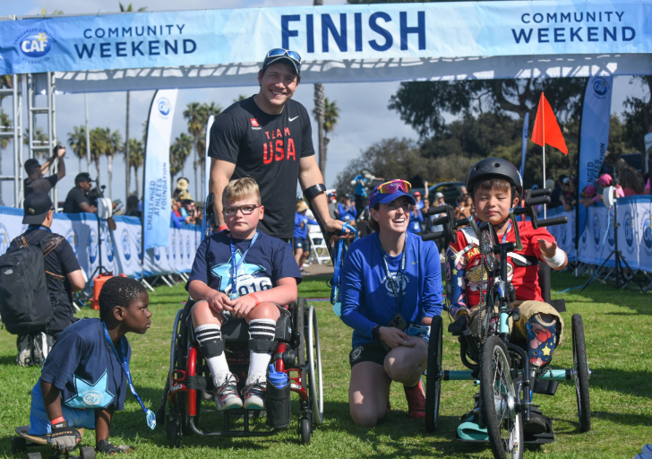 Paralympic alpine skier Andrew Kurka poses at a CAF Community Weekend finish line with young athletes using adaptive equipment on the grass.