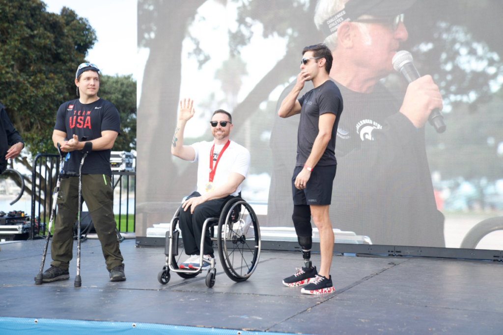 Andrew Kurka stands on stage at a CAF event alongside athletes and speakers during a program segment, with a large screen in the background.