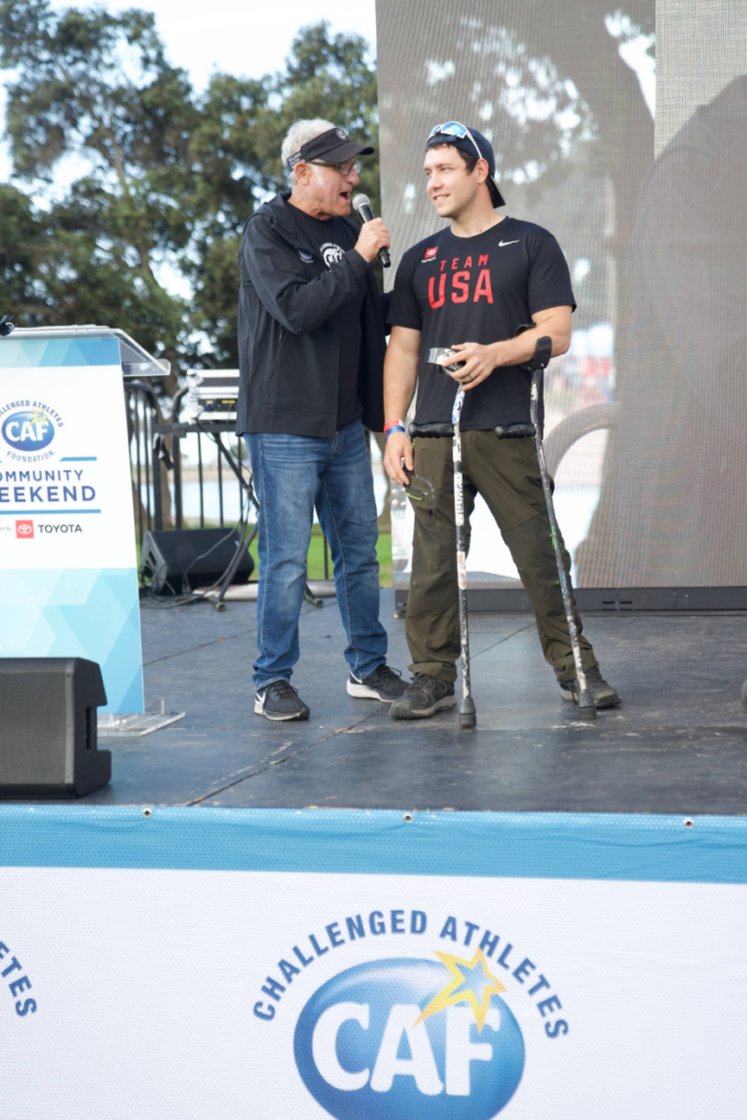 Andrew Kurka speaks on stage with a host at a CAF Community Weekend event, standing with forearm crutches beside a podium.
