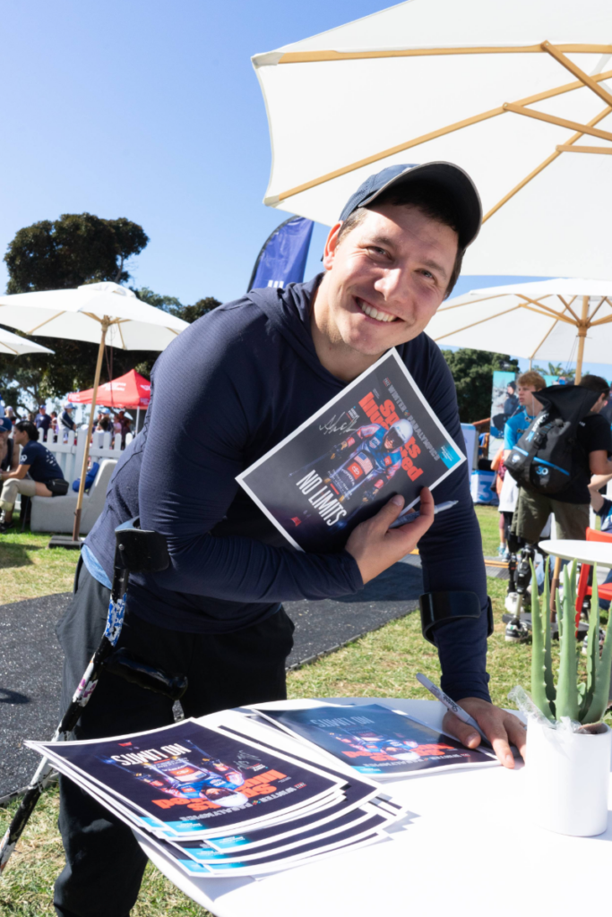 Andrew Kurka smiles while signing posters and greeting attendees at an outdoor CAF event, with umbrellas and participants in the background.