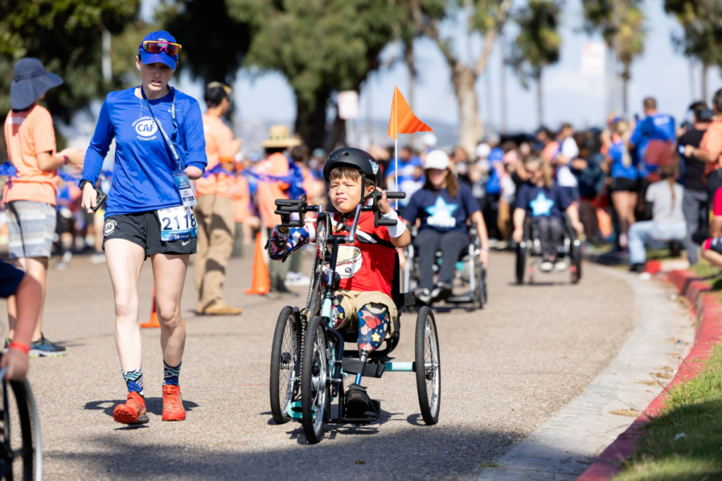 Dani Aravich runs alongside a young athlete using an adaptive handcycle during a CAF Community Weekend race, with participants and volunteers in the background.