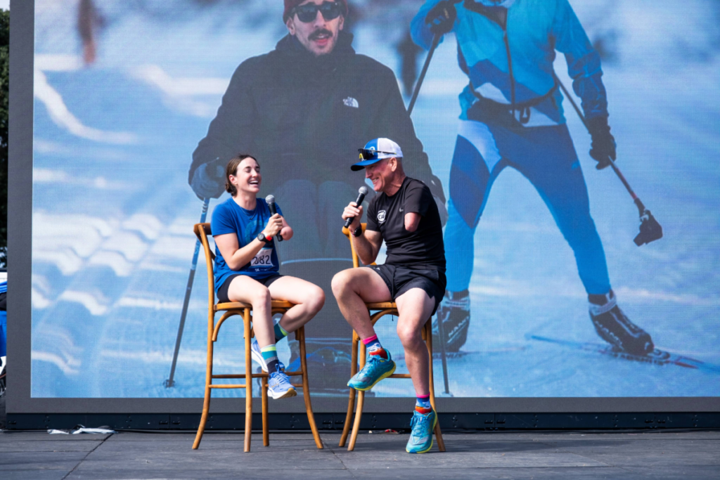 Dani Aravich speaks on stage during a live interview at a CAF event, seated with a host in front of a large screen showing Nordic skiing imagery.