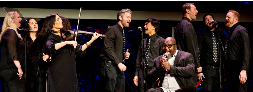 Live musical performance on stage at the Challenged Athletes Foundation Defying Limits Gala, featuring a vocalist and ensemble of singers alongside a violinist, with performers dressed in black under stage lighting.
