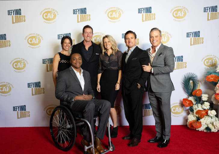 Group photo on the red carpet at the Challenged Athletes Foundation Defying Limits Gala, including an athlete with a physical disability seated in a wheelchair alongside event supporters, with CAF and Defying Limits logos in the backdrop.