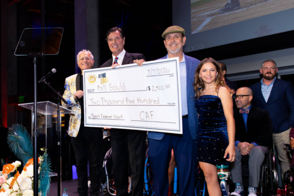 Group on stage at the Challenged Athletes Foundation Defying Limits Gala presenting a large grant check, with multiple individuals including athletes with physical disabilities and supporters gathered under stage lighting.