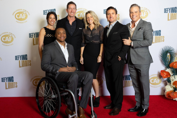 Group photo on the red carpet at the Challenged Athletes Foundation Defying Limits Gala, including an athlete with a physical disability seated in a wheelchair alongside event supporters, with CAF and Defying Limits logos in the backdrop.