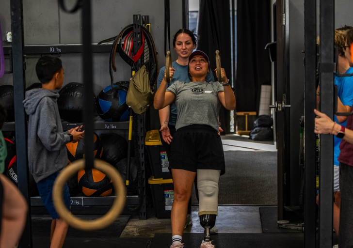 An athlete with a lower-limb prosthesis performs a ring row during a functional fitness clinic while a coach spots from behind, surrounded by other participants in a gym setting.