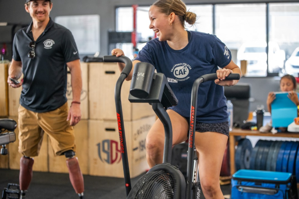 An athlete pedals an air bike while smiling during a functional fitness clinic as a coach observes nearby in a supportive gym environment.