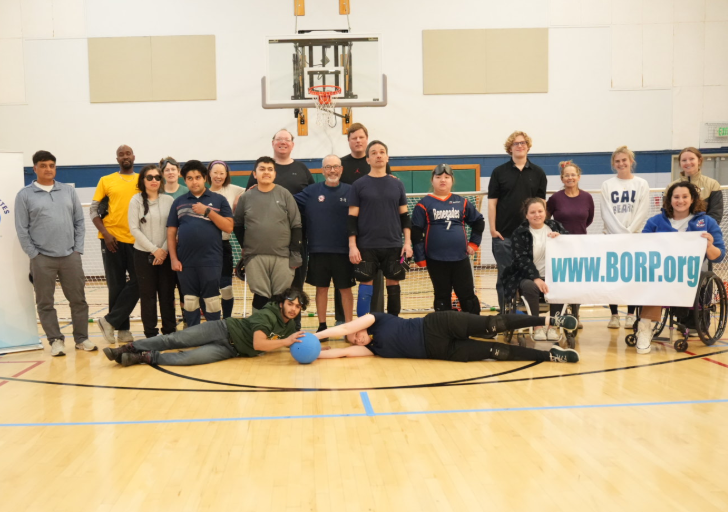 A group of goalball participants, coaches, and volunteers pose together on an indoor gym court during a CAF goalball clinic, standing behind goalball nets and holding a BORP banner, with two participants playfully lying on the floor in front holding a blue goalball.