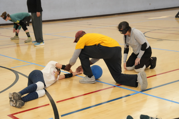 Two goalball participants dive across a gym floor reaching toward a blue goalball during a live drill, while a coach sits near the goal observing, with the goalball net stretched across the background.