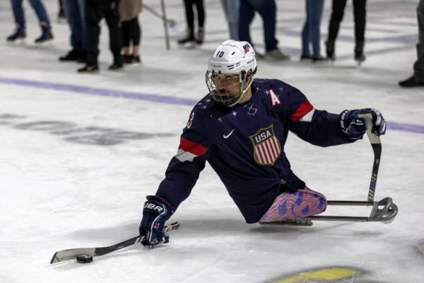 Athlete with a physical disability playing sled hockey on an indoor rink, wearing a Team USA jersey and helmet while handling the puck during a game.