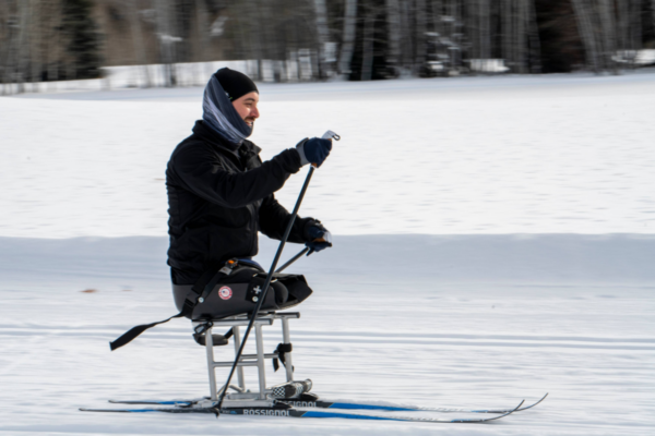 Athlete with a physical disability using a sit-ski to cross-country ski on a snow-covered trail, pushing forward with poles through a winter landscape.