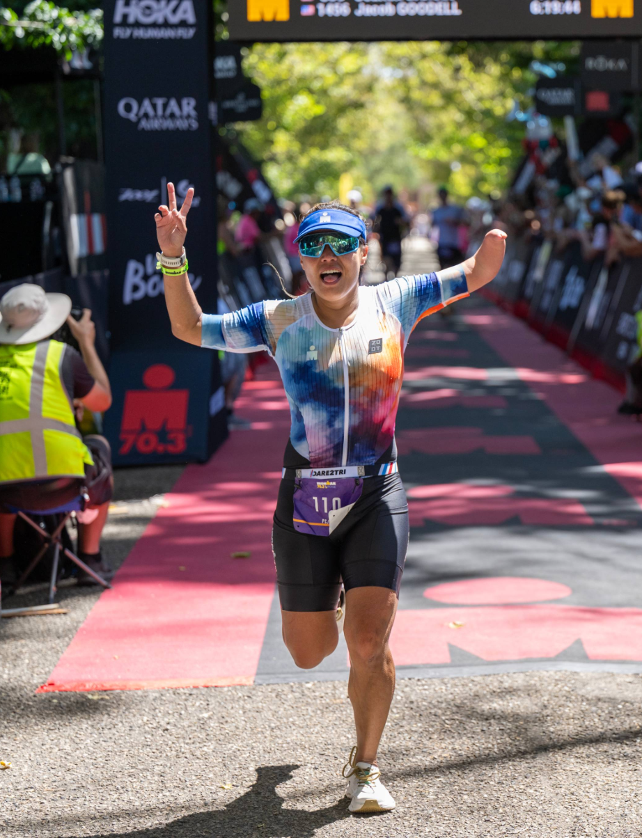 An athlete with a physical disability crosses the Ironman Boise finish line with arms raised in celebration, surrounded by spectators and race signage.
