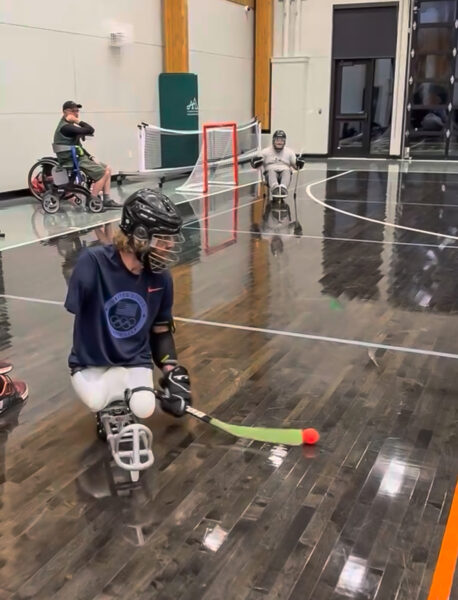 Athletes in wheelchairs playing floor hockey indoors, with a focus on one athlete in a navy blue shirt moving a ball with a green stick.