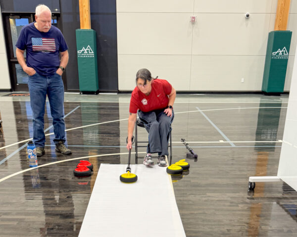 Person in a wheelchair engaged in curling, pushing a yellow stone on an indoor court, with another person observing.