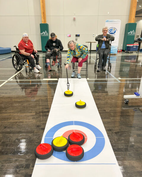 A group of four people in wheelchairs playing indoor curling, with stones of red and yellow on the target.