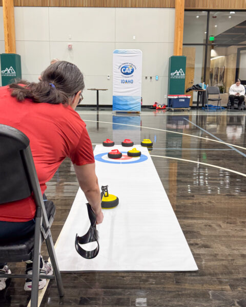 Person in red shirt playing indoor curling on a mat with red and yellow stones.