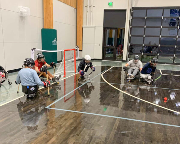 Individuals playing sled hockey indoors, using sleds and wearing helmets, near a goal and a red ball.