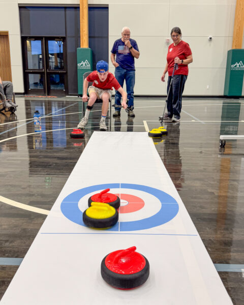 A young person pushes a curling stone on a mat in a gym, with two adults observing.