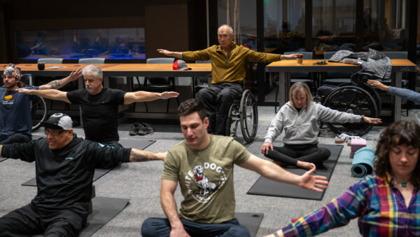 A diverse group of people doing yoga or stretching indoors, with some participants in wheelchairs.