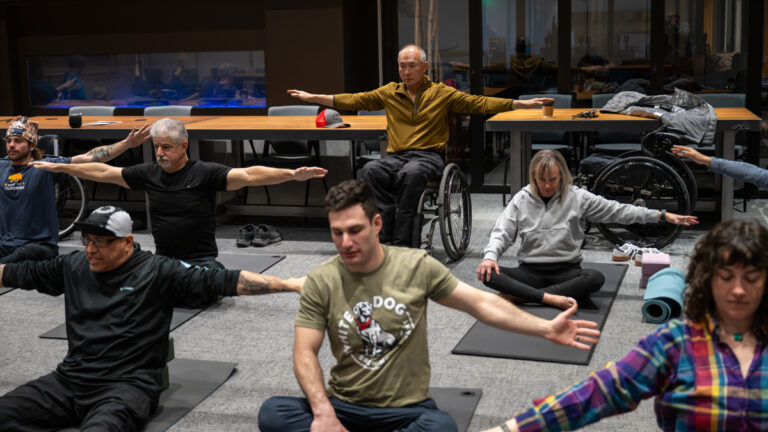 A diverse group of people doing yoga or stretching indoors, with some participants in wheelchairs.