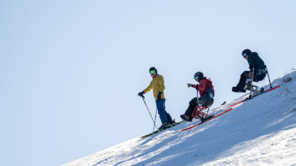 Three individuals on a snowy hillside, one standing on skis and two in sit skis.