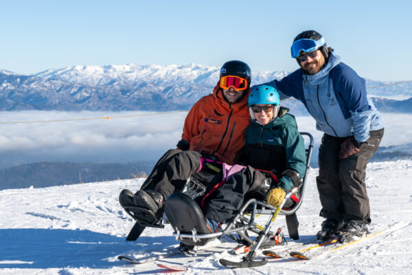 Three people on a snowy mountain, one using a sit-ski, with snow-capped peaks in the background.