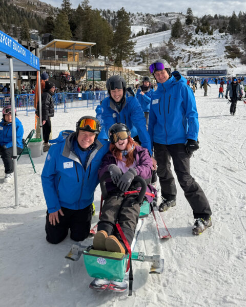 Group of people in winter sports gear, with a person on adaptive ski equipment in a snowy setting.