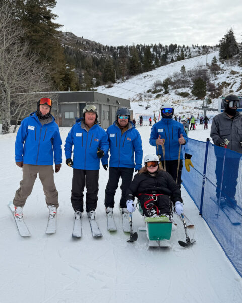 A group of people on a snowy ski slope, with four standing in blue jackets and one seated in an adaptive ski chair.