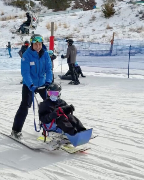 A person guides another in a blue adaptive ski on a snowy slope.