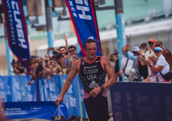 An athlete in a black USA racing kit crosses a finish chute at a paratriathlon event, with spectators cheering behind blue barricades and event flags lining the course.