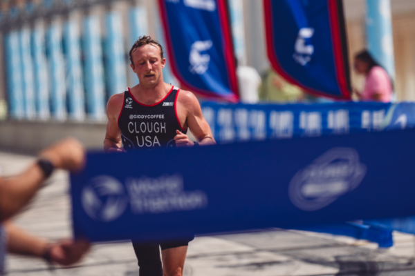 An athlete in a black USA racing kit crosses a finish chute at a paratriathlon event, with spectators cheering behind blue barricades and event flags lining the course.