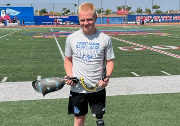 A student athlete stands at midfield on a football field holding a prosthetic running blade, wearing a championship team T‑shirt.