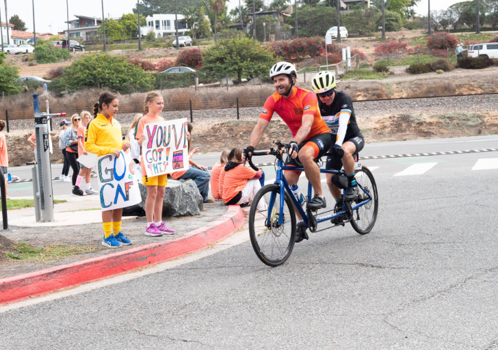 Cyclists ride a tandem bike during a charity ride while children cheer on the sidelines holding supportive signs for participants.