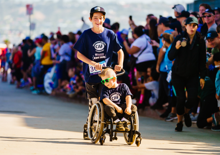 Brantley Volpel, a young athlete with spina bifida, smiles while participating in the Challenged Athletes Foundation Kids Run in his wheelchair with another participant guiding behind him.